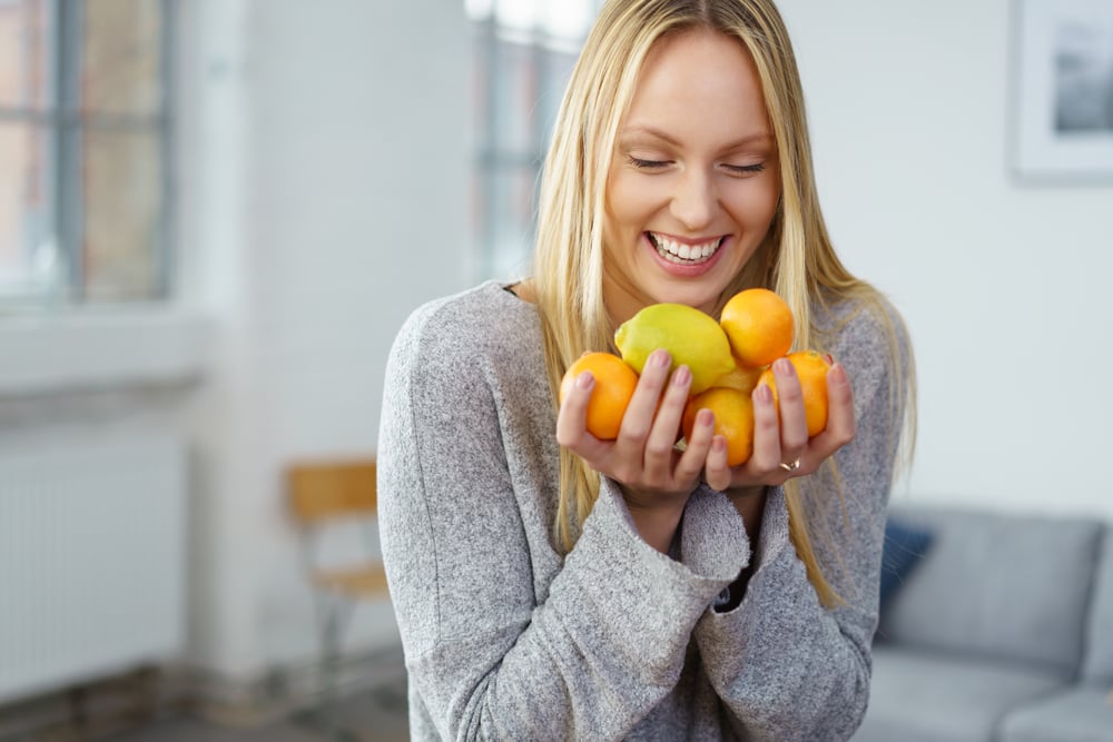 Happy young woman with a handful of fresh citrus fruit, lemons and oranges, standing in her living room grinning at the camera in a healthy diet concept